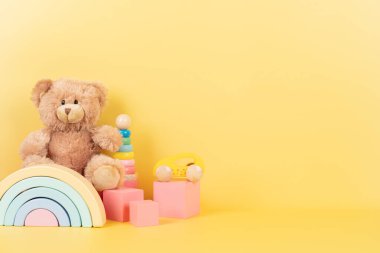 Educational kids toys collection. Teddy bear, wooden rainbow pink cubes and colorful balls on yellow background. Front view. clipart