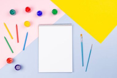 Yaratıcı sanat geçmişi sağlar. Suluboya, fırçalar, kalemler, pastel pembe, mavi, sarı arkaplan üzerine açık sarmal kağıt defter. Üst manzara, düz uzanma.