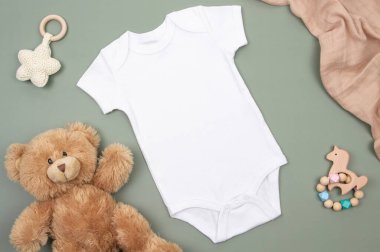 Beyaz çocuk tulumunun modeli ve beyaz ve kahverengi pamuklu yeşil arka planda çocuk oyuncakları. Yeni doğan bebekler için cinsiyet ayrımı olmayan kıyafetler. Üst görünüm.