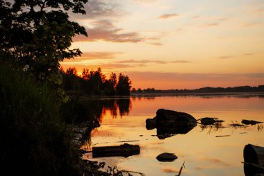 Akşam nehir kenarında, gökyüzünü kırmızımsı renklere boyayan bir gün batımı. Taşlar ve ağaçlar nehirde yatıyor..