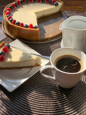 Güneşli bir günde masada meyvelerle süslenmiş bir fincan kahve ve ev yapımı peynirli kek.