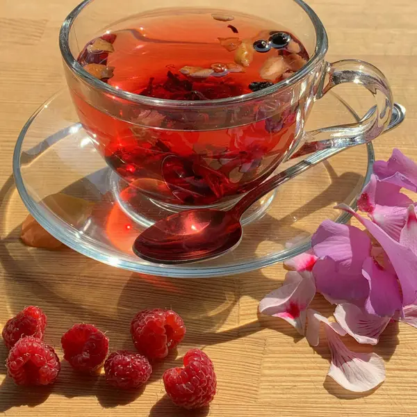 A cup of berry tea on a wooden table with flower petals and raspberries