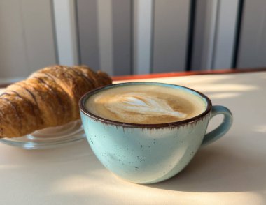 Kapuçino bardağının yakın çekimi ve masada bir kruvasan.