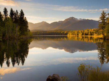 Sakin göl kıyıları, durgun sularda yükselen ağaçların ve uzak dağların yansımaları.