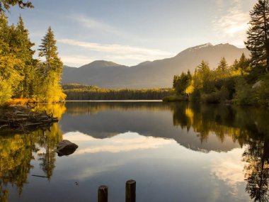 Sakin göl kıyıları, durgun sularda yükselen ağaçların ve uzak dağların yansımaları.
