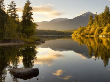 Sakin göl kıyıları, durgun sularda yükselen ağaçların ve uzak dağların yansımaları.