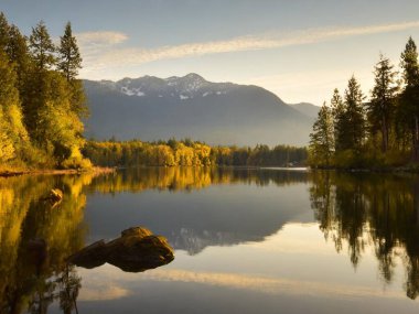 Sakin göl kıyıları, durgun sularda yükselen ağaçların ve uzak dağların yansımaları.