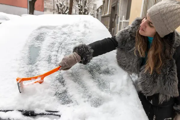 Young  woman using a snow brush to remove snow from her car\'s windshield. Concept of winter weather, car maintenance, and being prepared for snowy conditions. Transportation concept