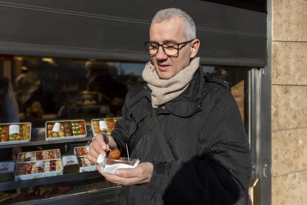 Middle age grey-haired man with black glasses and black jacket and beige scarf eating sweets on the street.