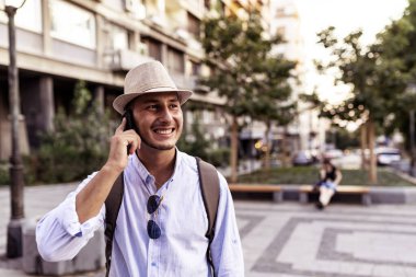 Yakışıklı İyi Giyimli Genç Adam Şapka, Güneş Gözlüğü ve Sırt Çantası Şehir Merkezinde Ayakta ve Cep Telefonu Üzerinden Talking