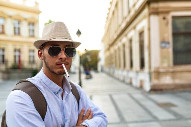 Yakışıklı İyi Giyimli Genç Adam Şapka, Güneş Gözlüğü ve Sırt Çantası Şehir Merkezinde Ayakta ve Sigara İçme