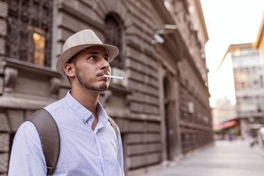 Yakışıklı İyi Giyimli Genç Adam Şapka, Güneş Gözlüğü ve Sırt Çantası Şehir Merkezinde Ayakta ve Sigara İçme