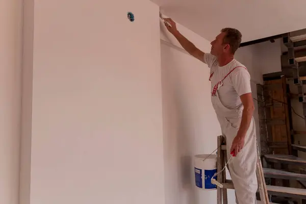 Male repairman painter paints the ceiling and wall of a house with a brush. Paint is applied to the wall by a professional worker.