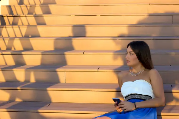 Mavi elbiseli, akıllı telefonunu kullanıp merdivenlerde oturan genç ve çekici bir kadın.
