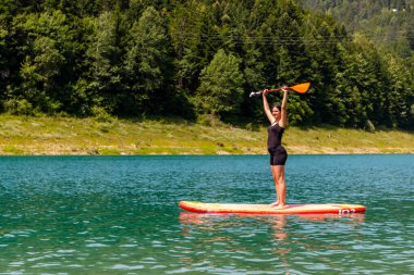 Genç bir kadın, parlak bir gülümseme ve neşeyle başının üstünde bir kürek tutarken gölde bir kürek tahtası üzerinde duruyor..