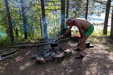 Ormandaki kamp ateşinde yemek pişiren orta yaşlı bir adam..