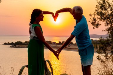 Mutlu yaşlı çift, denizde gün doğumunda elleri ve vücutlarıyla kalp şekli oluşturuyorlar.