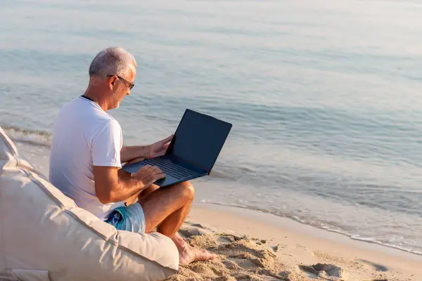 Sahilde oturmuş dizüstü bilgisayarı üzerinde çalışan günlük kıyafetli bir erkek turist. 