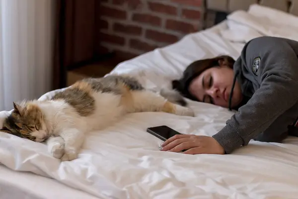 Elinde telefon olan genç bir kız ve yatakta huzur içinde uyuyan bir kedi.