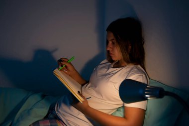 Genç bir kız, odasında yatağında uzanıyor ve gece yatağının başucundaki lambanın ışığıyla bazı notlar yazıyor. Genç kız odasında dinleniyor ve yazıyor..