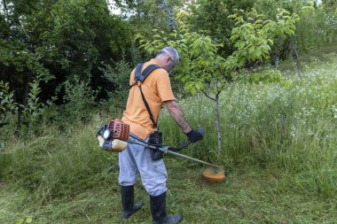 Koruyucu giysiler içinde olgun bir adam ve makasla eldivenler. Uzun çimleri biçmek ve yabani otları biçmek. Seçmeli olarak odaklan