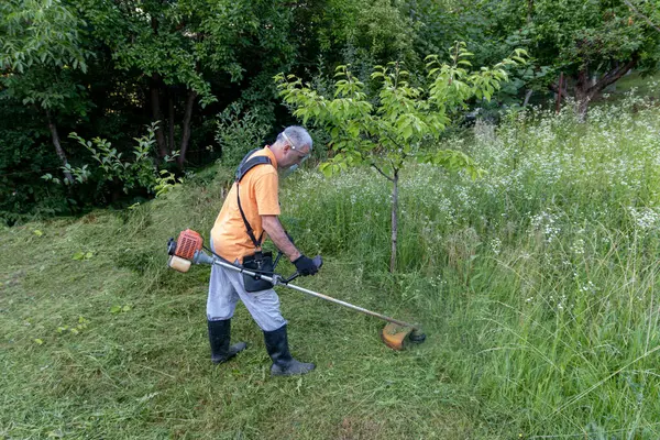 stock image A mature man in protective clothing, and gloves with a trimmer. Mowing tall grass and the weeds with a trimmer. Selectively focus