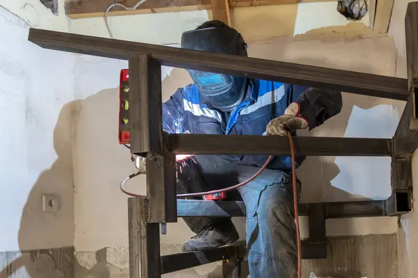 stock image A mature man welder in blue uniform, welding  metal with a arc welding machine at the construction site. An experienced man welding a seam on a spiral staircase at a construction site.