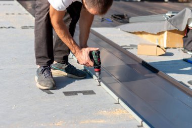 Roofer çatıda metal levha monte eder. Kalaycı işçi, kablosuz matkap vidaları kullanarak yeni metal levha çatısı kuruyor