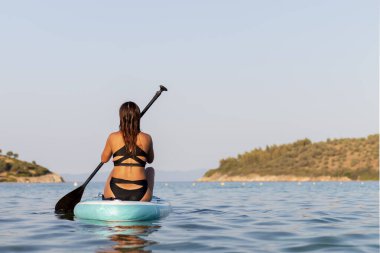 Kürek tahtasında duran ve kürek çeken genç bayan sörfçünün vücut arkası görüntüsü. Deniz kenarında yaz tatili boyunca aktif yaşam tarzı