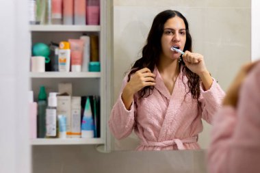 Gülümseyen bir kadının portresi dişlerini fırçalıyor ve banyodaki aynaya bakıyor. Ağız temizliği, sağlıklı dişler ve bakım. Sabah rutini. Kişisel temizlik.