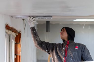 Apartman tavanını tek başına yenilemek için alçı kullanan genç bir erkek. Adam bir spatulayla tamir işi yapıyor. Apartman geliştirme ya da restorasyon işleri.