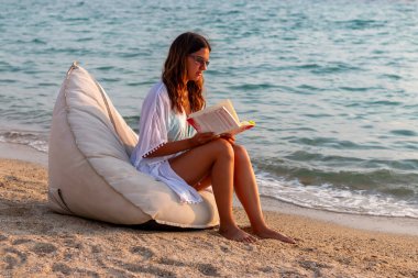 Güzel bir kadın gevşiyor ve koltukta kitap okuyor. Plaj tatili, dinlenme ve rahatlama kavramı..