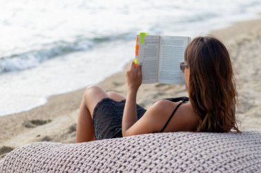 Kitap okuyan, kumsalda dinlenen, plaj yatağında oturan, fasulye koltuğu olan çekici bir kadın. Tatil konseptinde dinlen.. 