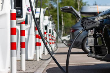 Yeşil çevredeki elektrikli araç şarj istasyonları. Araç şarj ediliyor. Batarya EV aracı fişe takıldı ve yenilenebilir enerji jenerasyonlarından elektrik alıyor güneş paneli, rüzgar türbini.