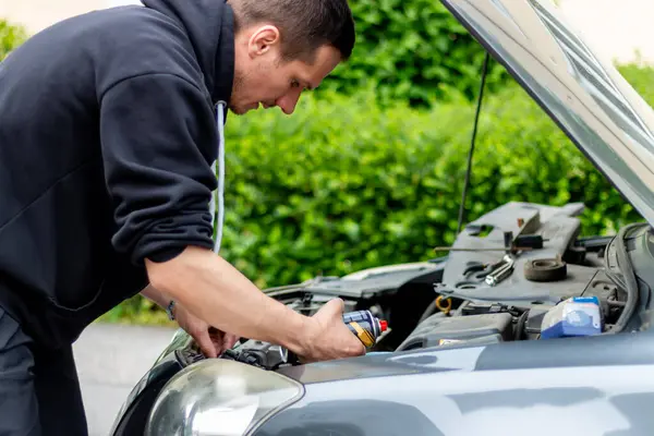 Genç bir adam paslı vidayı kimyasal bir maddeyle püskürtüp yağlıyor. Tamircinin eli açık kaputun altında çalışıyor..
