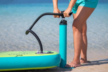 Genç ve güzel bir kadın deniz altyapısı olan plajda sörf tahtasını şişiriyor. Genç kız güneşli bir günde eğlenceye hazırlanıyor..