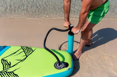 Olgun bir adam plajda deniz altyapısı olan kürek tahtasını şişiriyor. Güneşli bir günde son sınıf öğrencisi bir erkek eğlenceye hazırlanıyor. Kapat..