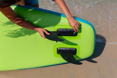 SUP tahtasının direksiyon yüzgeci. Genç bir kadın SUP tahtasının altındaki üsse yüzgeç sokuyor. Bir omurga montajı.