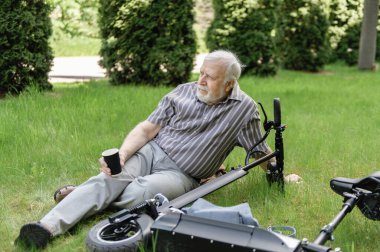 Olumlu, yaşlı bir adam parkta yürüyüşe çıktı, durdu ve elektrikli bir scooterın yanındaki çimlerin üzerinde dinlenmek için uzandı. Yaz, güneşli bir gün. Yaşlıların sağlıklı yaşam tarzı.