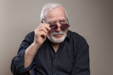 Charismatic elderly gray-haired man in sunglasses and gray shirt posing for camera in studio, looking to the side with a smile on light background