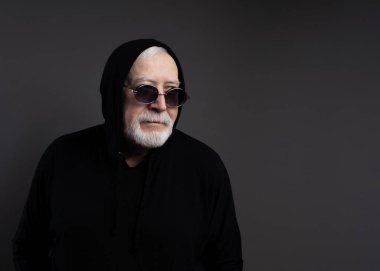 Charismatic elderly gray-haired man in sports black suit, hood and sunglasses, studio, gray background, place for text