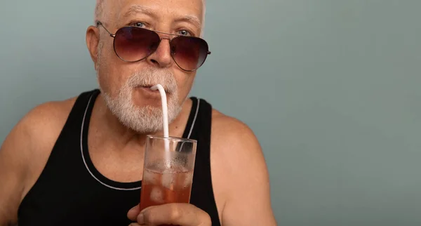 A charismatic elderly man with sunglasses and a black T -shirt holds an icy drink, an interesting facial expression, a light background, a place for text