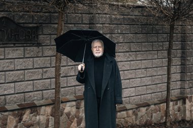 Şık, karizmatik, şemsiyeli yaşlı bir adam dışarıda çitin yanındaki yağmurda, yağmurlu havada bekliyor.