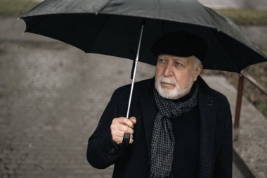 Siyah ceketli, şemsiyeli yaşlı bir adam yağmurda merdivenlerde duruyor, yağmurlu bir havada.