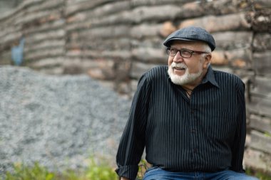 Gözlüklü ve şapkalı gülümseyen yaşlı bir adam ahşap bir çitin arkasında oturuyor, yan tarafa bakıyor, yarım boy portre, sokak, metin için yer