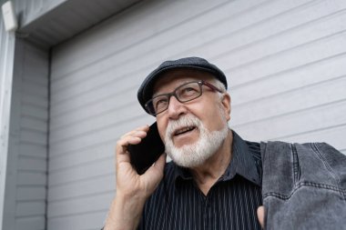 Gözlüklü karizmatik yaşlı bir adam ve gri bir garaj kapısının arkasında duran bir şapka, telefonla konuşuyor, yatay format.