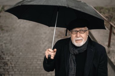 Siyah ceketli, şemsiyeli yaşlı bir adam yağmurda merdivenlerde duruyor, yağmurlu bir havada.
