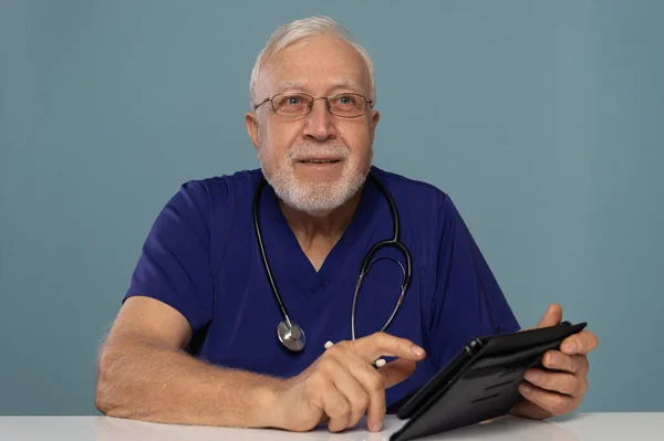 A gray-haired, elderly doctor, wearing glasses and a stethoscope, is telling a client something in his office, pointing at a clipboard