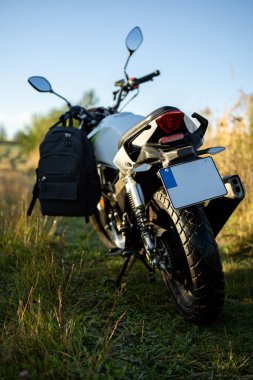 A motorcycle stands parked on a grassy area, with a black backpack hanging from its side. The sun shines brightly, creating a vibrant, outdoor scene.