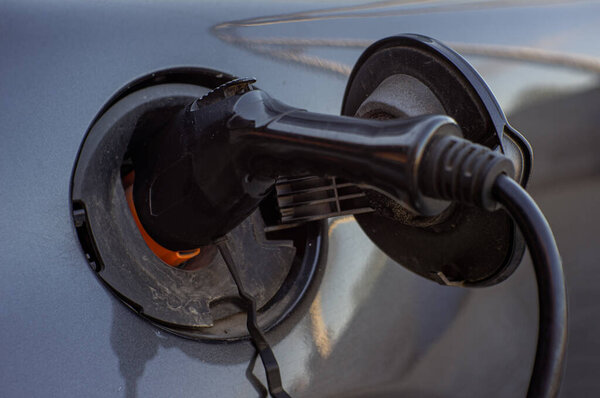 An electric vehicle is connected to a charging station, with a charging cable plugged into the car. The background shows a typical urban environment during late afternoon.